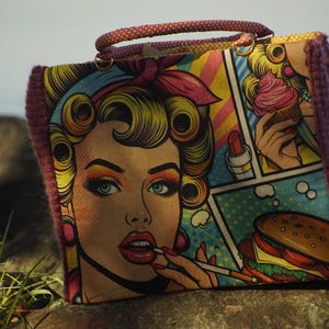 Può includere: Borsa in stile pop art con un'illustrazione a fumetti di una donna con capelli biondi, occhi blu e rossetto rosso. La borsa ha un bordo in maglia viola e un manico a pois. Il fumetto include immagini di un hamburger e gelato.