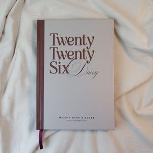 May include: A closed diary with the text "Twenty Twenty Six Diary" in a dark brown font. The cover is light gray with a dark brown spine and a burgundy ribbon bookmark. The diary includes weekly pages and notes.