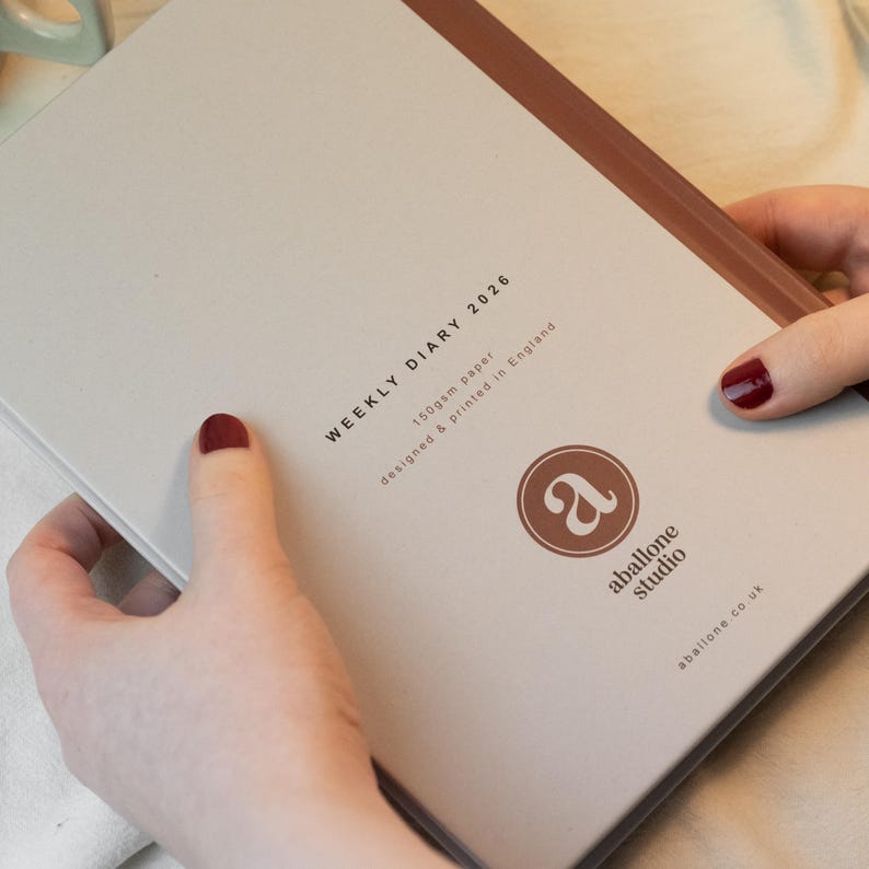May include: A light beige weekly diary for 2026, with a brown spine. The cover features the text "WEEKLY DIARY 2026", "150gsm paper", and "designed & printed in England". The diary has a logo that says "aballone studio".