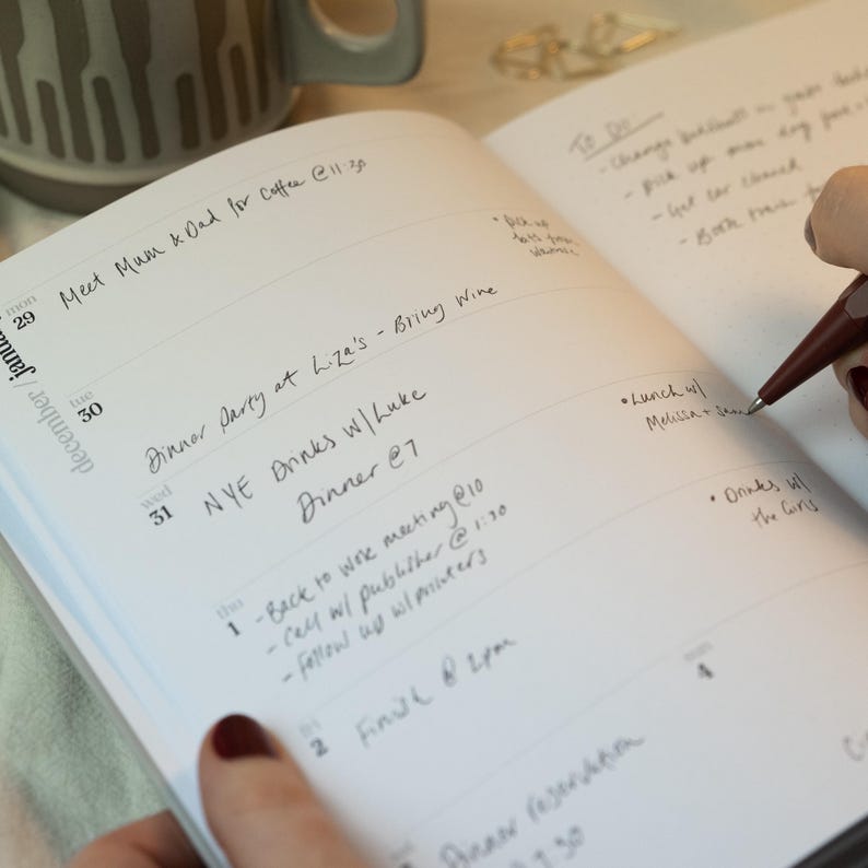 May include: An open, lined planner with handwritten entries for December and January. The planner includes daily schedules, appointments, and to-do lists. A pen is poised over the page, ready to add more notes. A mug is visible in the background.