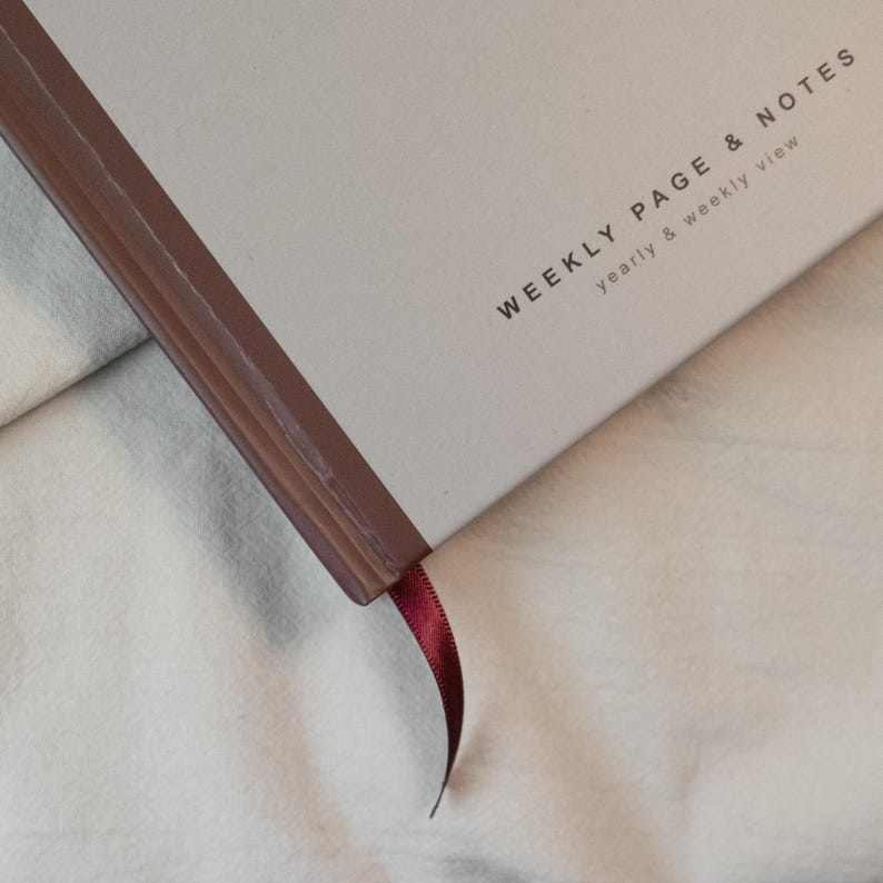 May include: A close-up of a notebook with the text "WEEKLY PAGE & NOTES" and "yearly & weekly view" printed on the cover. The notebook has a burgundy spine and a matching ribbon bookmark.