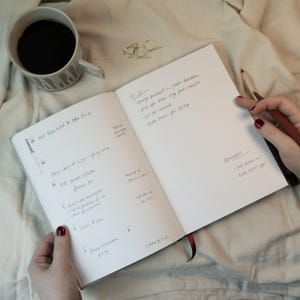 May include: An open notebook with handwritten notes, a cup of coffee, and a pen. The notebook's pages display a to-do list and reminders. The coffee cup has a gray and white pattern. The scene is set on a soft, white surface.
