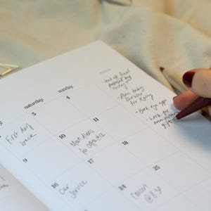 May include: A close-up of a white calendar with handwritten notes in black ink. The calendar is open to the month of October, with the days of the week and dates clearly visible. A hand with a red pen is writing on the calendar.