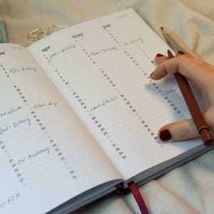 May include: Open planner with dates for April, May, and June 2020. Handwritten notes mark birthdays and events. A person's hand holds a red pen, poised over the June calendar. A red ribbon bookmark is visible.