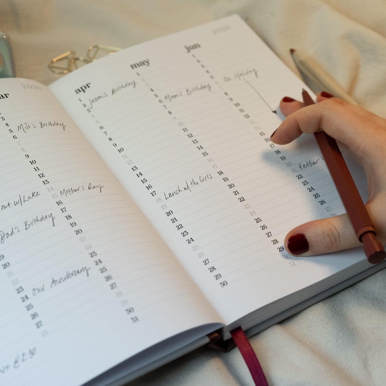 May include: Open planner with dates for April, May, and June 2020. Handwritten notes mark birthdays and events. A person's hand holds a red pen, poised over the June calendar. A red ribbon bookmark is visible.