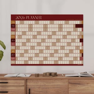 May include: A 2026 planner with a burgundy border and the text "2026 Planner" at the top. The calendar grid is in a neutral beige colour, with each month labelled vertically on the left side. The planner is displayed on a wooden surface.