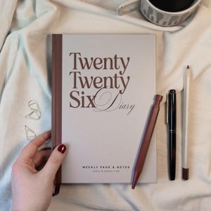 May include: A light gray diary with the text "Twenty Twenty Six Diary" in brown script. A brown pen, a black pen, and a white pencil are next to the diary. Two silver paper clips and a mug are also in the image.