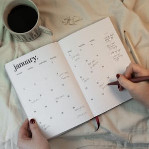 May include: An open planner with the month of January displayed, featuring handwritten notes on the calendar dates. A cup of coffee and a pencil are next to the planner. The planner is open to a page with the word "January" at the top.