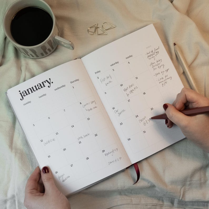May include: An open planner with the month of January displayed, featuring handwritten notes on the calendar dates. A cup of coffee and a pencil are next to the planner. The planner is open to a page with the word "January" at the top.