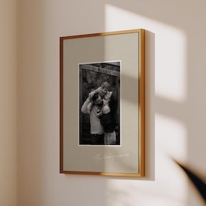 May include: A framed black and white family photograph. The image is set within a light beige mount and a thin, gold-coloured frame. The words "the lucky ones" are written in cursive at the bottom of the mount.