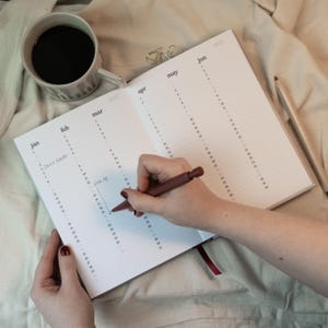 May include: An open planner with the months of the year printed on the pages. A person is writing in the planner with a brown pen. A cup of coffee and a pencil are next to the planner.