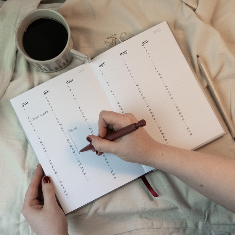 May include: An open planner with the months of the year printed on the pages. A person is writing in the planner with a brown pen. A cup of coffee and a pencil are next to the planner.
