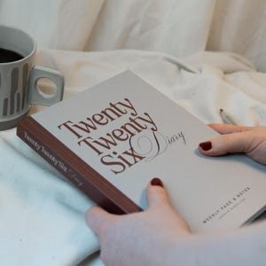 May include: A diary with the text "Twenty Twenty Six Diary" in brown lettering. The diary has a light gray cover and a brown spine. A mug of coffee is visible in the background.