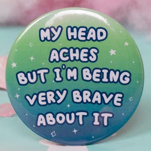 Peut inclure: Un bouton rond avec un dégradé de vert à bleu, portant l'inscription blanche "MY HEAD ACHES BUT I'M BEING VERY BRAVE ABOUT IT". Le fond pastel est orné de barbe à papa rose et d'étoiles décoratives.