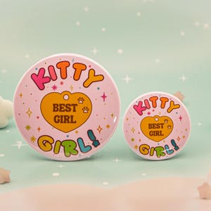 Op de afbeelding: Twee roze ronde buttons met de woorden "KITTY BEST GIRL" in kleurrijke tekst. De grotere button heeft een diameter van ongeveer 7,6 cm, de kleinere button ongeveer 3,8 cm. De buttons hebben een retro-ontwerp met een hart en pootafdrukken.