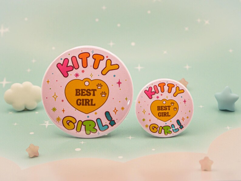 Op de afbeelding: Twee roze ronde buttons met de woorden "KITTY BEST GIRL" in kleurrijke tekst. De grotere button heeft een diameter van ongeveer 7,6 cm, de kleinere button ongeveer 3,8 cm. De buttons hebben een retro-ontwerp met een hart en pootafdrukken.