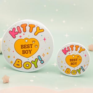 Op de afbeelding: Twee witte ronde buttons met de tekst "KITTY BEST BOY" in kleurrijke letters. Het ontwerp bevat een gele hart met pootafdrukken en sterren. E&eacute;n button is groter dan de andere.