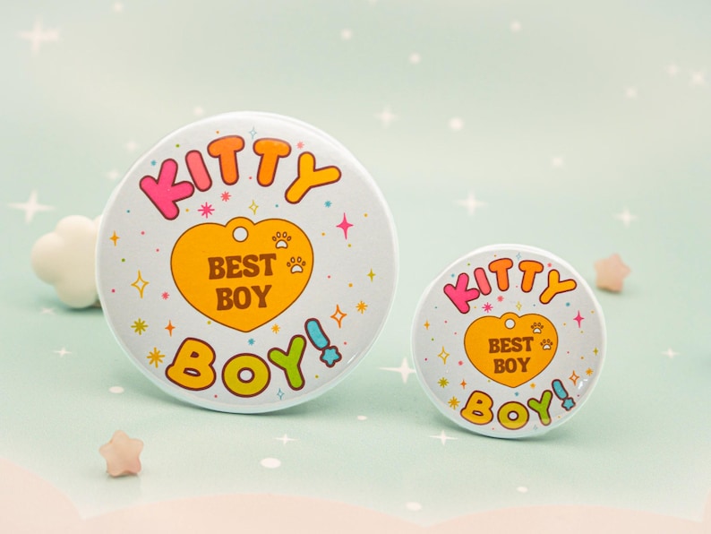 Op de afbeelding: Twee witte ronde buttons met de tekst "KITTY BEST BOY" in kleurrijke letters. Het ontwerp bevat een gele hart met pootafdrukken en sterren. E&eacute;n button is groter dan de andere.
