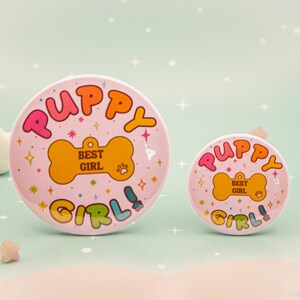 Op de afbeelding: Twee roze ronde buttons met de woorden "Puppy Girl!" in kleurrijke bubbelletters. De buttons hebben een botvormig label met "Best Girl" erop. De achtergrond is licht turquoise met sterren.
