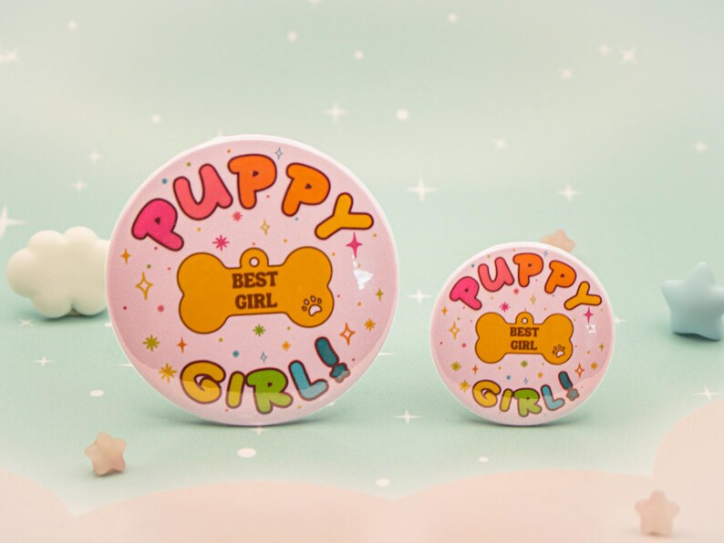 Op de afbeelding: Twee roze ronde buttons met de woorden "Puppy Girl!" in kleurrijke bubbelletters. De buttons hebben een botvormig label met "Best Girl" erop. De achtergrond is licht turquoise met sterren.