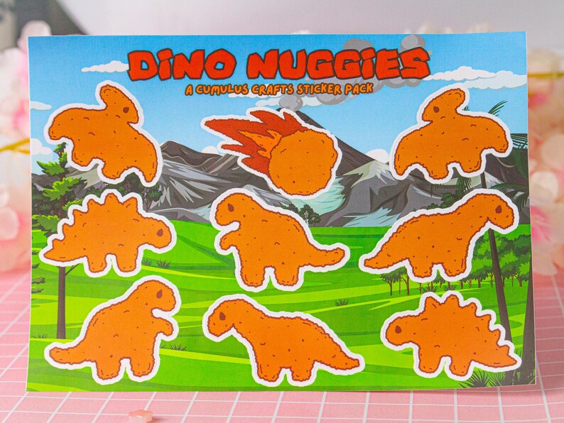 May include: A sticker pack featuring eight dinosaur-shaped chicken nuggets. The stickers are orange and have a cartoonish style. The text "Dino Nuggiges" and "A Cumulus Crafts Sticker Pack" is printed on the sticker sheet.