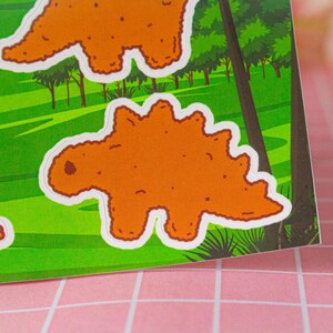 May include: A set of four orange dinosaur stickers with white outlines. The dinosaurs are in various poses, including standing, walking, and running. The stickers are on a green background with trees and bushes.