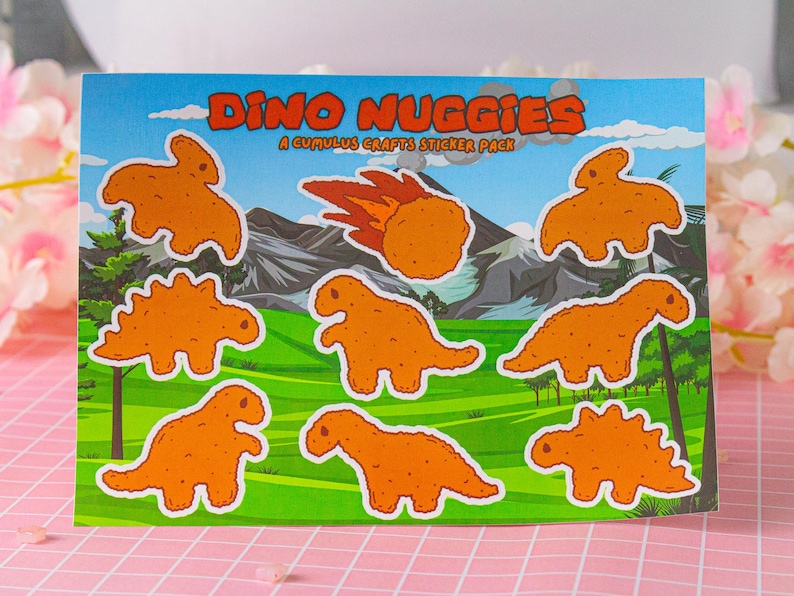 May include: A sheet of stickers featuring cartoon dinosaur-shaped chicken nuggets. The stickers are orange and yellow and have the text "Dino Nuggiges A Cumulus Crafts Sticker Pack".
