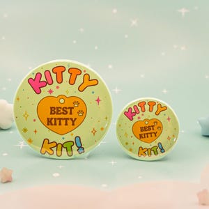 Op de afbeelding: Twee ronde, lichtgroene knopen met de tekst "KITTY" in roze en oranje, en "BEST KITTY" in een hartvorm. De knopen hebben een pootafdrukontwerp en het woord "KIT!" in blauw. De achtergrond is lichtblauw met sterren en decoratieve vormen.