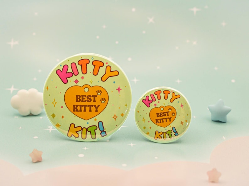 Op de afbeelding: Twee ronde, lichtgroene knopen met de tekst "KITTY" in roze en oranje, en "BEST KITTY" in een hartvorm. De knopen hebben een pootafdrukontwerp en het woord "KIT!" in blauw. De achtergrond is lichtblauw met sterren en decoratieve vormen.