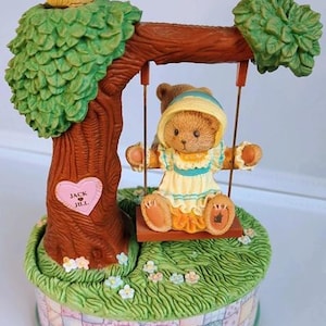 May include: A decorative figurine featuring a teddy bear on a swing under a tree. The bear wears a yellow and blue outfit. The base has a patchwork design with the words "Cherished Teddies". A blue bird sits atop the tree.