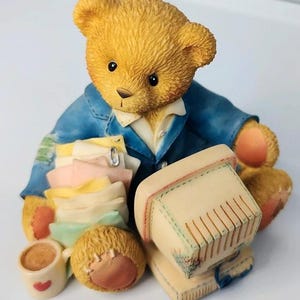 May include: A detailed figurine of a teddy bear wearing a blue jacket and holding a stack of papers. The bear has a computer, a cup of coffee with a heart, and a patch on its arm. The figurine is made of a light-colored material.