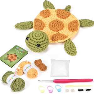 Pode incluir: Um kit de crochê de crochê com um porta-copos de tartaruga finalizado, com casco amarelo e marrom e cabeça e nadadeiras verdes. O kit inclui fio verde e amarelo, um saco de enchimento, uma agulha de crochê rosa, marcadores de pontos e um folheto.