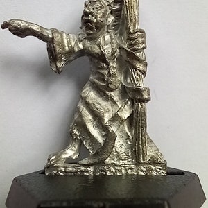 May include: A silver metal miniature figurine of a wizard holding a staff. The wizard is wearing a long robe and has a beard. The figurine is standing on a black base.