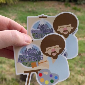 May include: Two stickers featuring a cartoon of Bob Ross painting. The top sticker says "Some Inspirational Bullsh*t" and the bottom sticker says "Happy Accidents". Both stickers have a mountain scene in the background and a paint palette with colorful paint.