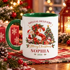 Könnte beinhalten: Ein weißer Keramikbecher mit grünem Henkel und Innenraum, verziert mit einem rot karierten Buchstaben S, dem Weihnachtsmann, einem Tannenbaum, einem Schneemann und Geschenken. Der Text lautet SPECIAL DELIVERY, Merry Christmas, SOPHIA und 2026.