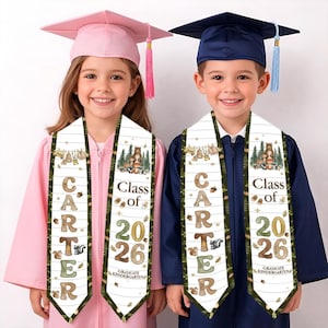 Könnte beinhalten: Zwei Kinder in Abschlusskleidung, jeweils mit einer Schärpe mit der Aufschrift "CARTER" und "Class of 2026". Die Schärpen sind mit Waldtier-Designs verziert. Ein Kind trägt eine rosa, das andere eine marineblaue Kappe und Robe.