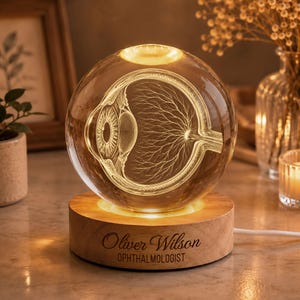 Könnte beinhalten: Eine klare Glaskugel mit einer internen, lasergravierten anatomischen Darstellung eines menschlichen Auges auf einem runden Holzsockel. Der Sockel ist mit dem Text "Oliver Wilson" und "OPHTHALMOLOGIST" in einer dunklen Schrift graviert.