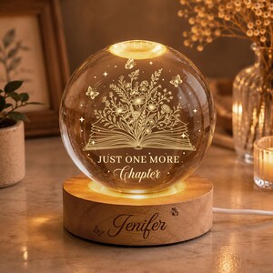 Könnte beinhalten: Ein kugelförmiges Glas-Nachtlicht mit einer Gravur eines offenen Buches, Blumen und Schmetterlingen. Der Text "JUST ONE MORE Chapter" ist in das Glas geätzt. Es steht auf einem runden Holzsockel, der mit dem Namen "Jenifer" graviert ist.