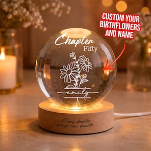 Puede incluir: Una esfera de vidrio transparente con diseño floral grabado, las palabras "Chapter Fifty" y un nombre. La esfera se asienta sobre una base de madera con el texto "Every chapter brings new growth." La esfera está iluminada con un brillo cálido.