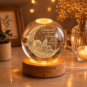 May include: A spherical glass night light on a round wooden base. The glass features an engraved crescent moon with a sleeping angel and the text: Carried for a moment, loved for a lifetime, Baby Emma, 2026. The wooden base is engraved: Forever in our hearts.