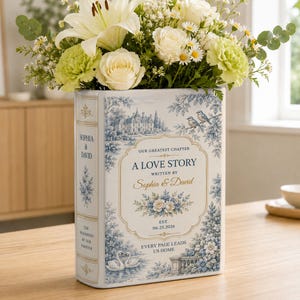 May include: A rectangular ceramic vase shaped like a book with blue and white toile-style illustrations of a manor, birds, and swans. The text reads: A LOVE STORY, WRITTEN BY Sophia & David, OUR GREATEST CHAPTER, EST. 06.25.2026, EVERY PAGE LEADS US HOME.