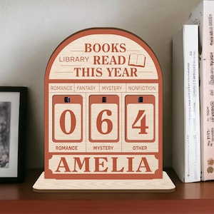 Könnte beinhalten: Holzregal-Display mit den Worten "BOOKS LIBRARY READ THIS YEAR". Es hat drei Zahlenanzeigen, die "064" für gelesene Bücher anzeigen. Der Name "AMELIA" wird unten angezeigt. Das Display steht auf einem Regal mit Büchern.