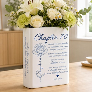 May include: A white rectangular ceramic vase featuring blue text and a rose illustration. The front displays the text Chapter 70, a rose, and a sentimental poem. The name Sophia is written along the rose stem. The side reads New chapter, new dreams, same beautiful you. Est. 1955 is printed at the bottom.