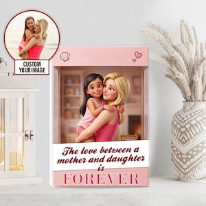 Może przedstawiać: Różowe pudełko dekoracyjne z kreskówkowym wizerunkiem matki i córki obejmujących się. Pudełko zawiera napis "The love between a mother and daughter is FOREVER". Mniejszy obrazek matki i córki znajduje się w lewym górnym rogu.