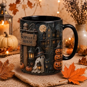 Könnte beinhalten: Ein schwarzer Keramikbecher mit einer detaillierten Halloween-Szene, die ein Spukhaus, einen Geist mit Laterne, eine schwarze Katze und drei gestapelte Kürbisse zeigt. Ein hölzernes Schild trägt die erhabene Aufschrift SOPHIA'S HOUSE.