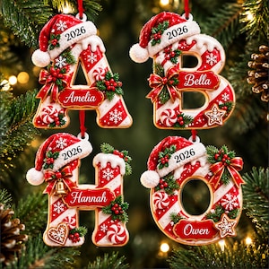 Könnte beinhalten: Vier Weihnachtsbaumanhänger in Form der Buchstaben A, B, H und O. Jeder Buchstabe hat ein rot-weißes Zuckerstangenmuster, eine Weihnachtsmütze mit der Jahreszahl 2026, grüne Stechpalmen, rote Beeren, weiße Schneeflocken und ein Namensschild.