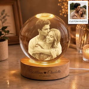 Könnte beinhalten: Eine kugelförmige Glaslampe mit einem lasergravierten Bild eines sich umarmenden Paares. Die Glaskugel ruht auf einem runden Holzsockel, der mit den Namen William und Charlotte sowie dem Datum 07.16.2026 graviert und von unten beleuchtet ist.