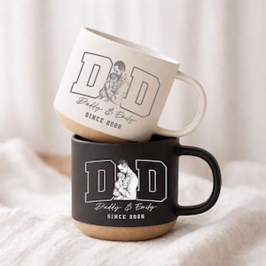 Könnte beinhalten: Zwei Keramikbecher, einer weiß und einer schwarz, übereinander gestapelt. Jeder Becher zeigt das Wort "DAD" in fetten Buchstaben, eine Skizze eines Vaters, der ein Kind hält, und den Text "Daddy & Emily SINCE 2025" und "SINCE 2026".