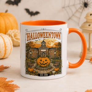Könnte beinhalten: Ein weißer Keramikbecher mit orangefarbenem Innenraum und Henkel, der eine grafische Illustration eines Gebäudes mit dem Text HALLOWEENTOWN und EST. 1998 zeigt. Das Design umfasst einen großen Kürbiskopf, Bäume und einen Vollmond im Hintergrund.