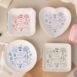 Peut inclure: Quatre petits plats à bijoux en céramique blanche, de formes carrée, en cœur et ronde, avec un bord doré. Les plats présentent des dessins illustrés et du texte, dont "it's giving wifey", "Henry + Rose" et "future Mrs. Taylor."